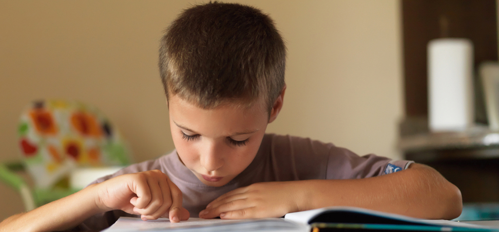 a boy sitting with a book open looking down and reading and pointing with his finger at the book. 