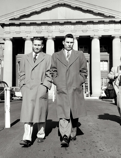two young men walk side-by-side, each dressed in a topcoat, circa 1950s