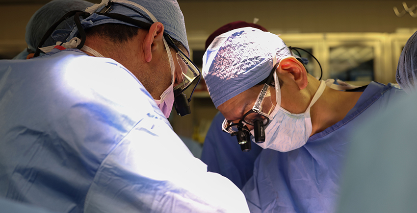 Photo of surgeons in scrubs and masks operating on an unseen patient. Only one is seen head-on. He wears attachments on his glasses to magnify what he's seeing.