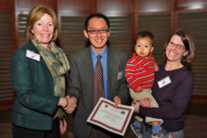 Roy Ahn (center), joined by his wife, Amy Hintzman (right), and son, Charlie, accepts a Shore Fellowship certificate from Dean for Academic and Clinical Affairs Nancy Tarbell. Photo by Steve Gilbert.