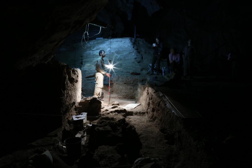 Man with lamp stands in a cave, an excavated skull in front of him