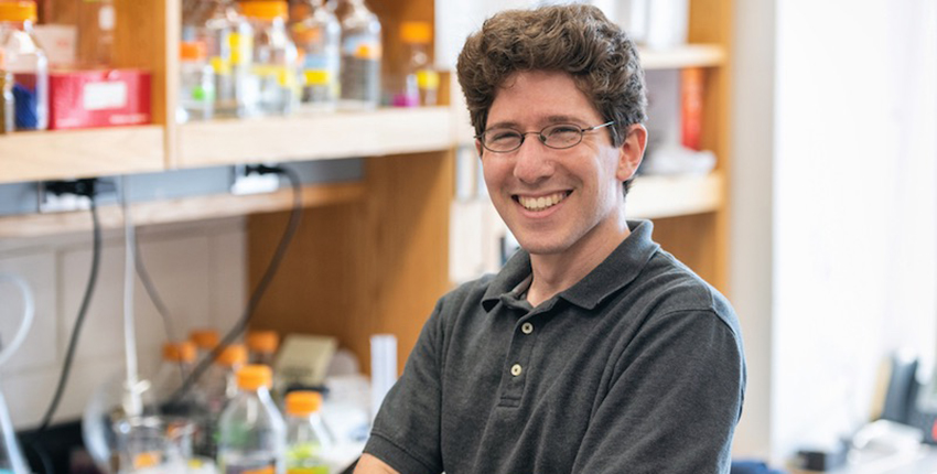 Young white man with curly brown hair standing in lab smiling