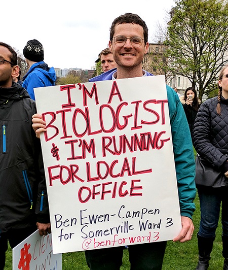 Young man stands in crowd outdoors with sign saying "I'm a biologist and I'm running for local office"