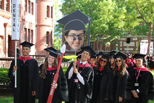 Eight grads in regalia carrying a standard that reads Dental, a four-foot Harvard toothbrush, and a cardboard cut out with a classmate's head.