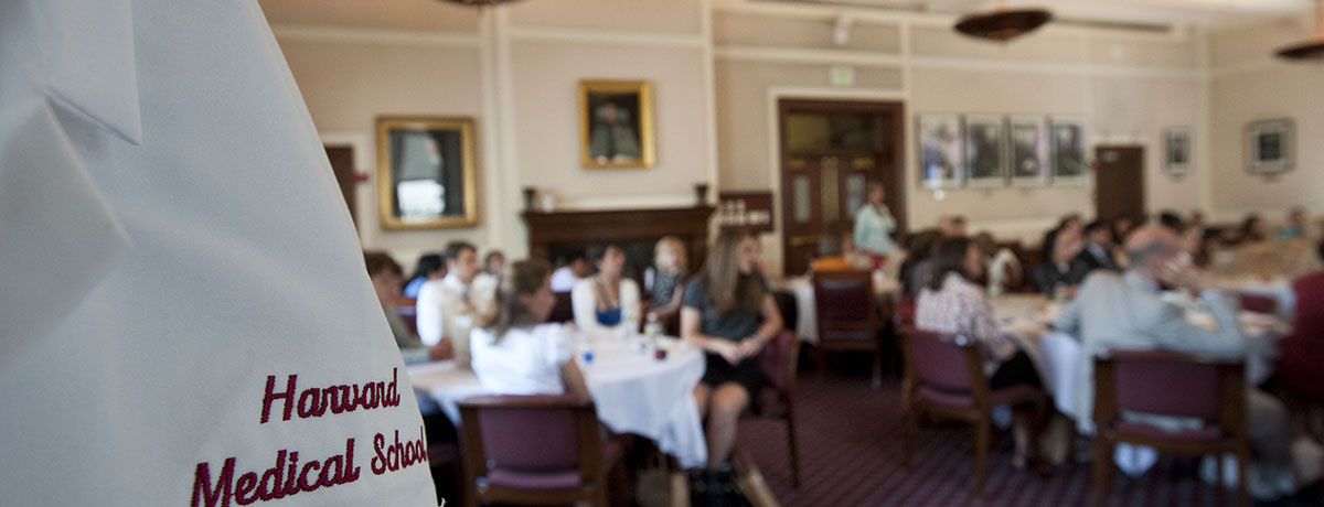 Photo of white coat with "Harvard Medical School" embroidered on it and students sitting at tables in the background