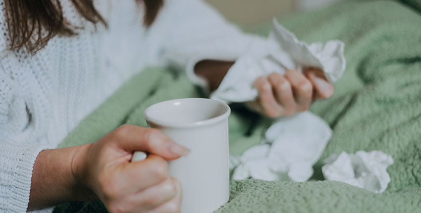 a person sitting in bed under a blanket, holding tea and surrounded by crumpled facial tissues