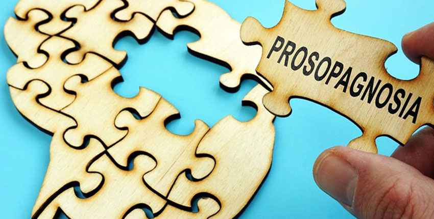 A hand holds a puzzle piece that reads "prosopagnosia" above a puzzle in the shape of a human brain