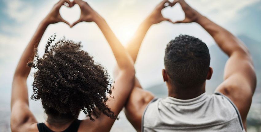 Photo of a Black young man and young woman making heart signs with their hands, backs facing the camera