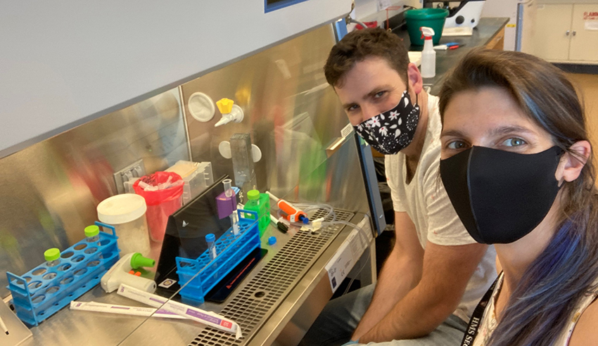 Two people in masks smile beside a hooded lab enclosure