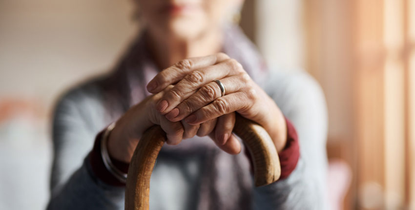An out-of-focus person sits with their hands on top of a cane in focus in front of them