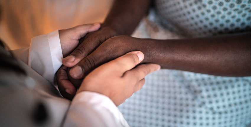 Image of a white coated doctor's hands holding the hands of a black patient wearing a hospital Johnny. No faces.