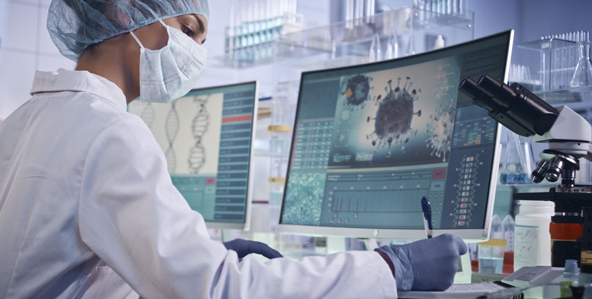 A scientist sits writing notes in front of a computer monitor showing a COVID-19 virus