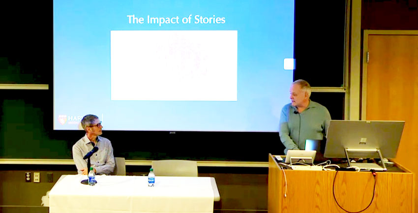 Zoom screen grab of the lecture with Foster seated at a table and Baer speaking from a podium inside a classroom.