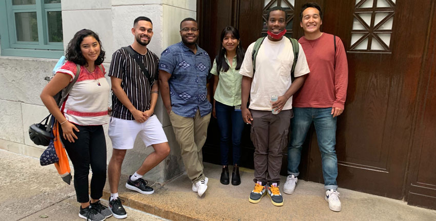 Six students pose for a photo against a stone building and a wooden door