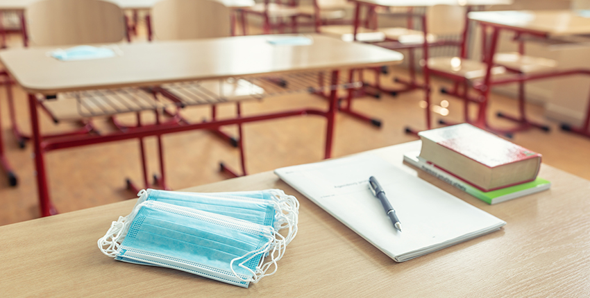 A stack of surgical masks, a pad of paper and pen, and a pile of books sit on a table in an empty classroom