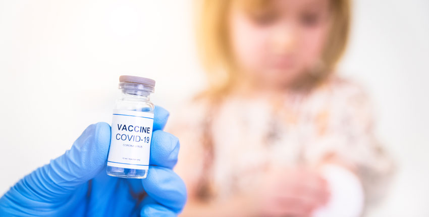 Blue-gloved hand holding COVID vaccine vial and blurry child in background	 