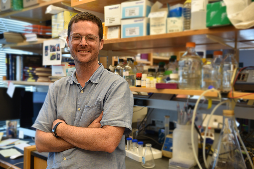 Young white man stands smiling with arms crossed in front of lab bench full of bottles