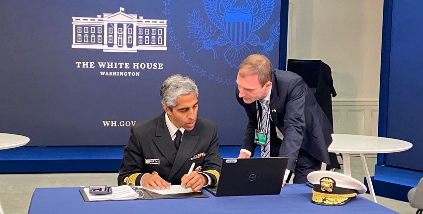 Murthy in uniform seated at table looking at laptop with Beckman to right leaning in; a backdrop behind them reads The White House