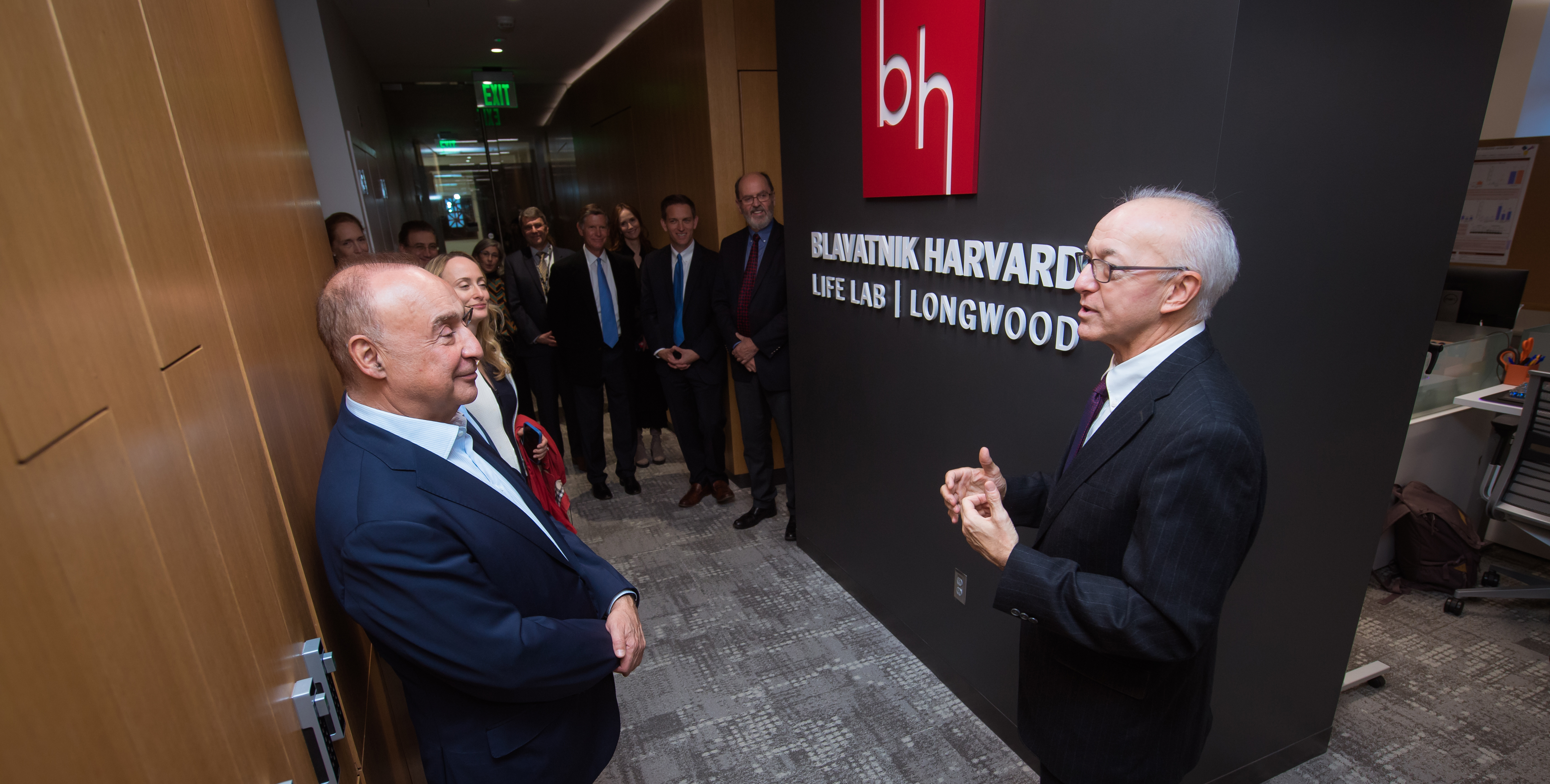 Two men in suits stand in front of a sign with a red square logo with letter B and H above white type on black background that reads Blavatnik Harvard Life Lab Longwood