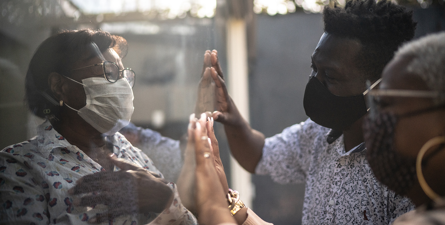 A woman on one side of a window faces a man and a woman on the other side, all three wearing protective masks.