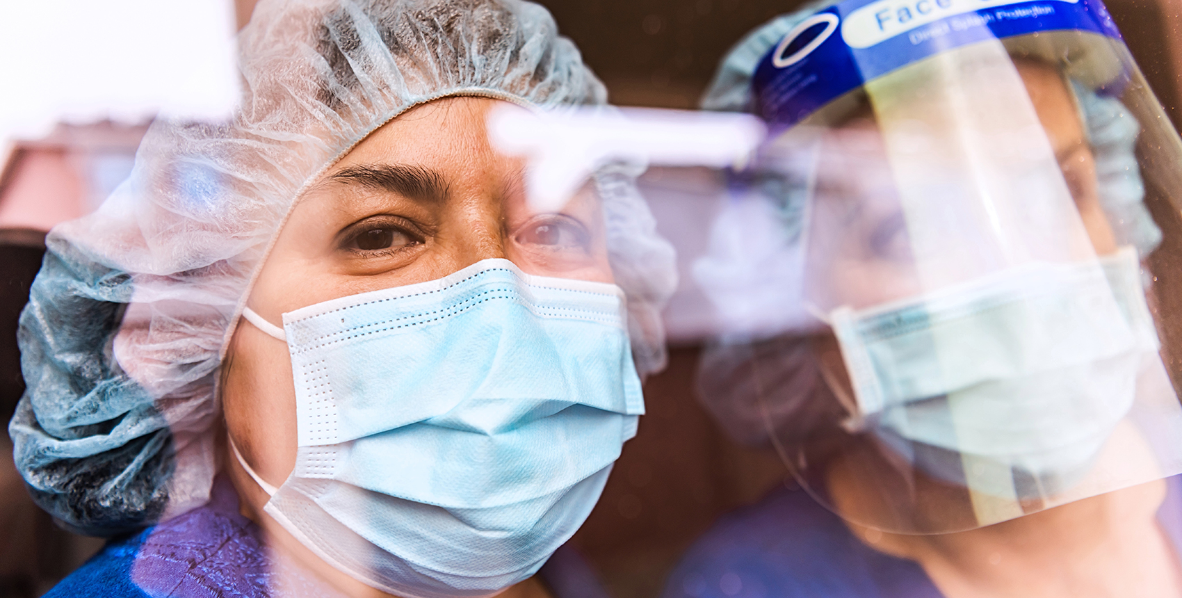 The faces of two people in medical scrubs, protective masks, and head gear seen through a reflection-covered window.