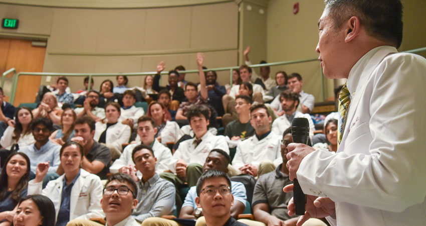 Sideview of Chang in white coat holding mic while speaking to audience of students, many in white coats
