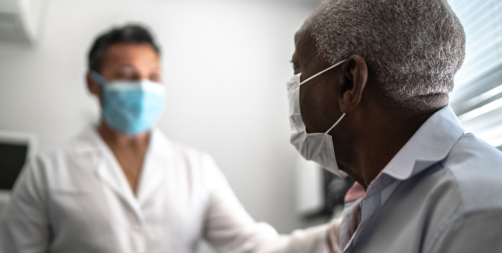 A dark-haired man in a white lab coat sits in a brightly lit room with a gray-haired man in a dress shirt.