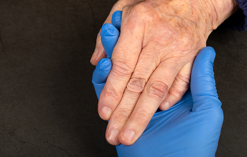 A hand in a blue latex glove holds the ungloved hand of an older white person