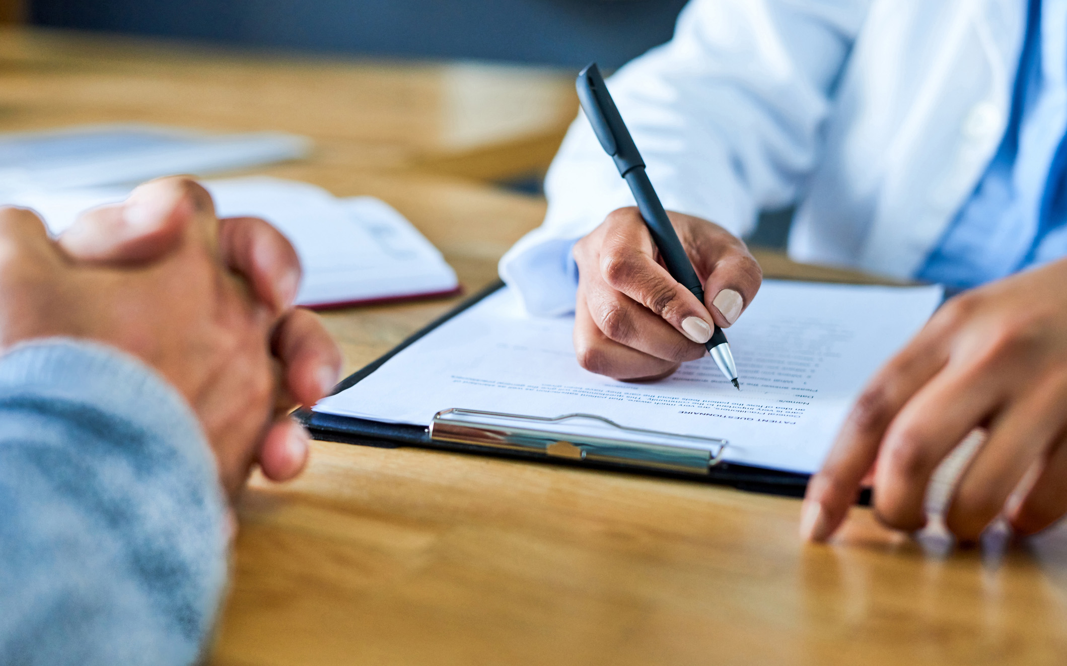 Photograph of doctor's hand writing on a form across from patient with hands folded