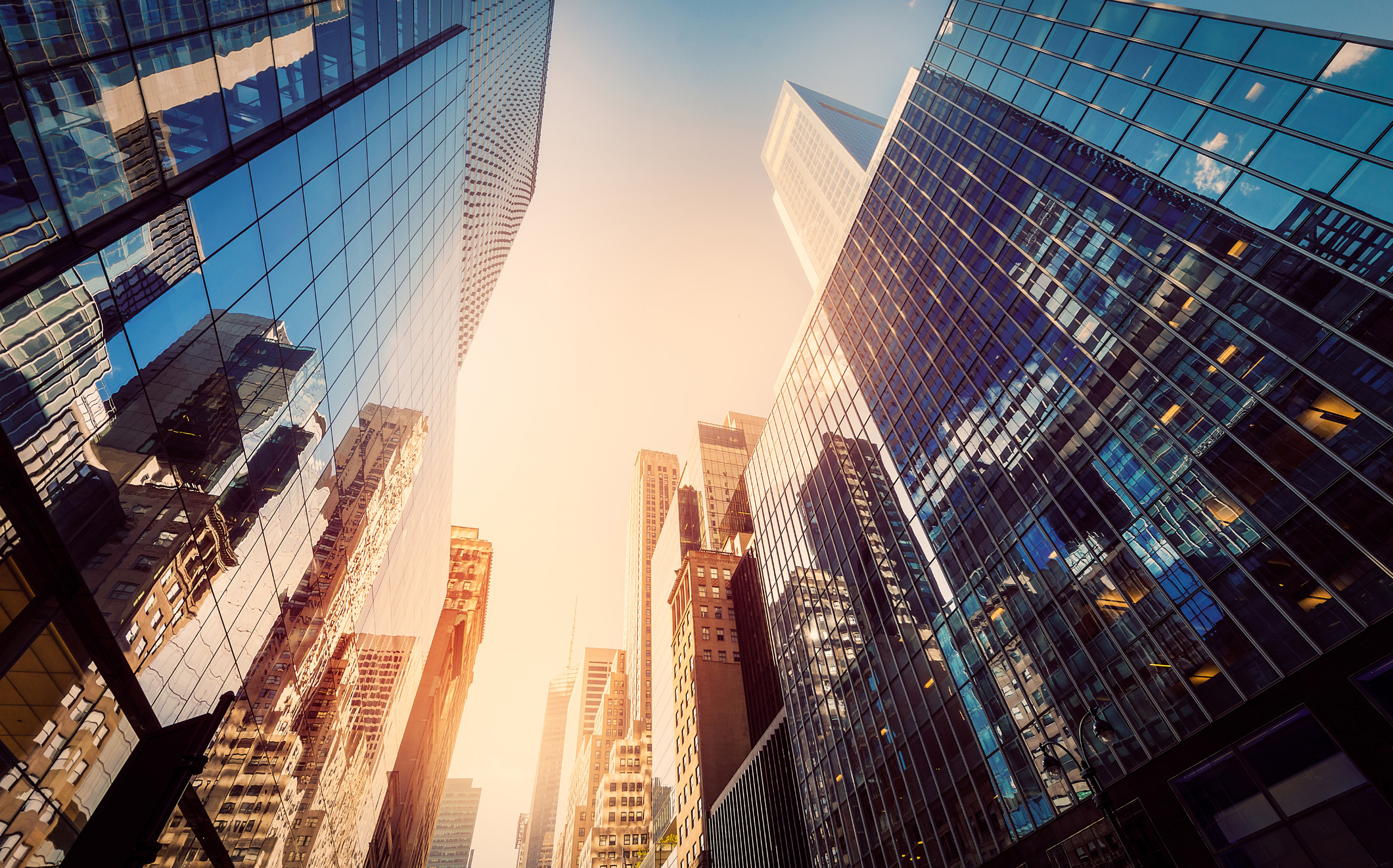 Photograph of tall office buildings, taken from the street, looking up to a sunny sky and tops of skyscrapers