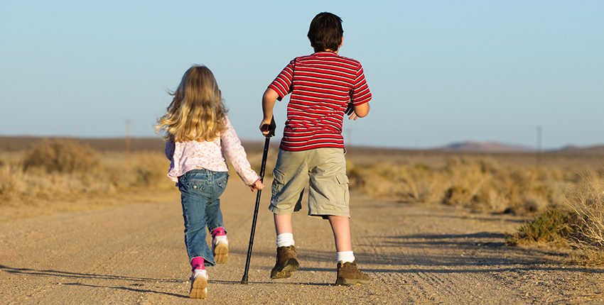 Two children - one girl unassisted and one boy with a crutch - skip down a dirt road