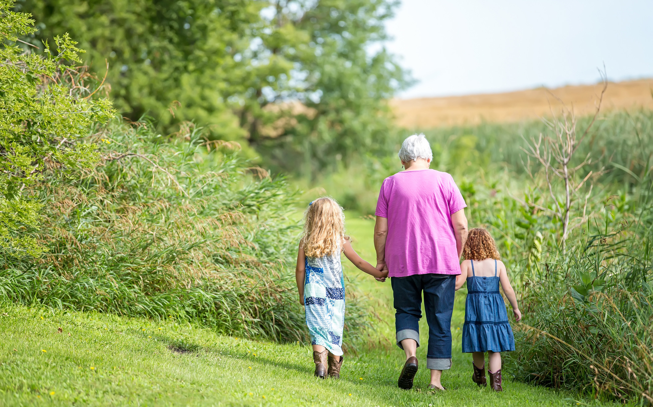 Woman in pink top walking, holding hands with two little girls, backs to camera, in a garden