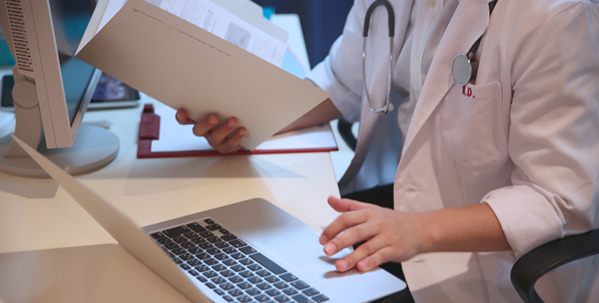 Photo of physician holding a pen and a notebook and typing on a computer keyboard