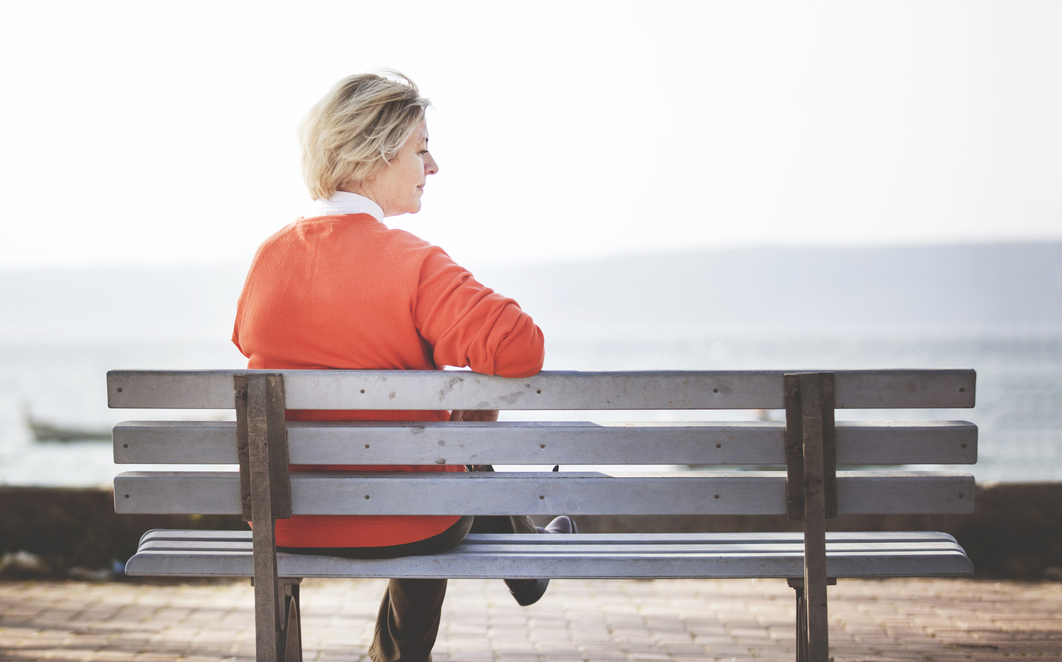 Woman with light hair and orange sweater sits alone on a bench overlooking water