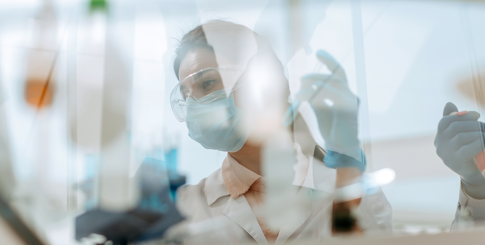 A woman in goggles mask and lab coat working at a laboratory table seen through the reflections and refractions on a safety glass window.