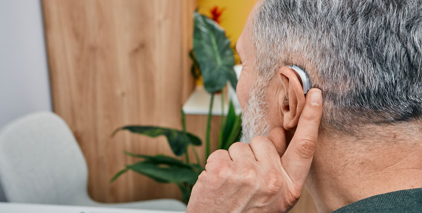 back of the head of a white grey-haired man holding a finger to his ear that has a hearing aid