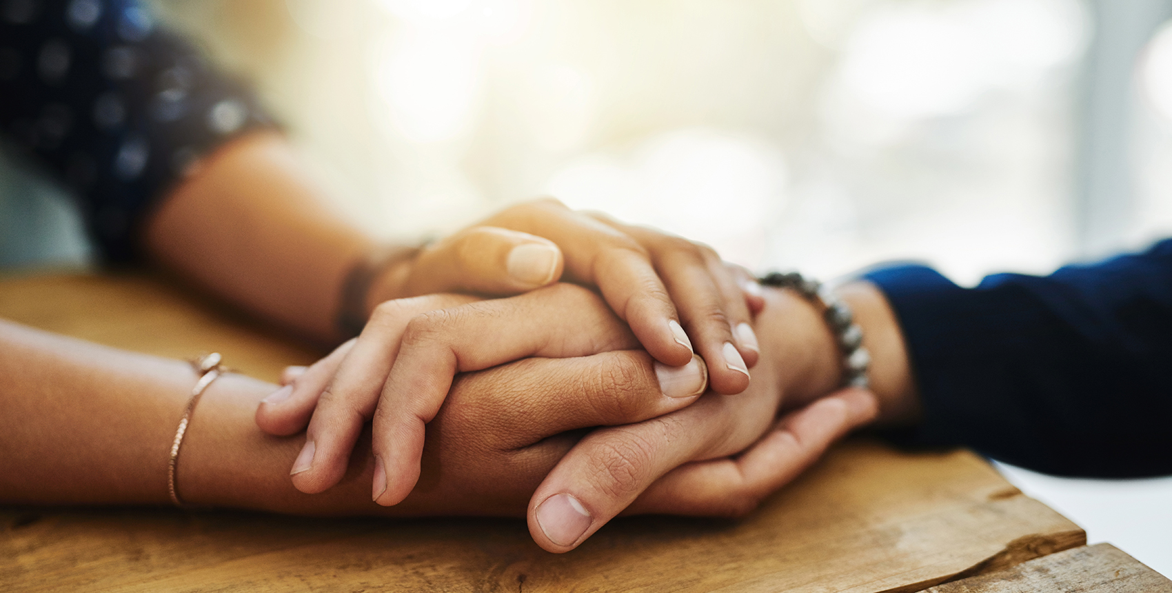 Closeup of two people’s hands clasped together on a table in a comforting manner.
