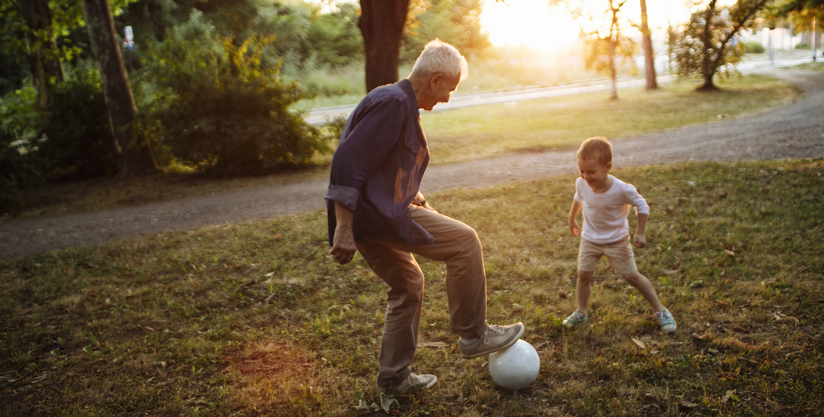 Caught in the middle of a one-on-one soccer game, a wite-haired man stands with his foot on a soccer ball, a small child faces him, poised to take the ball.
