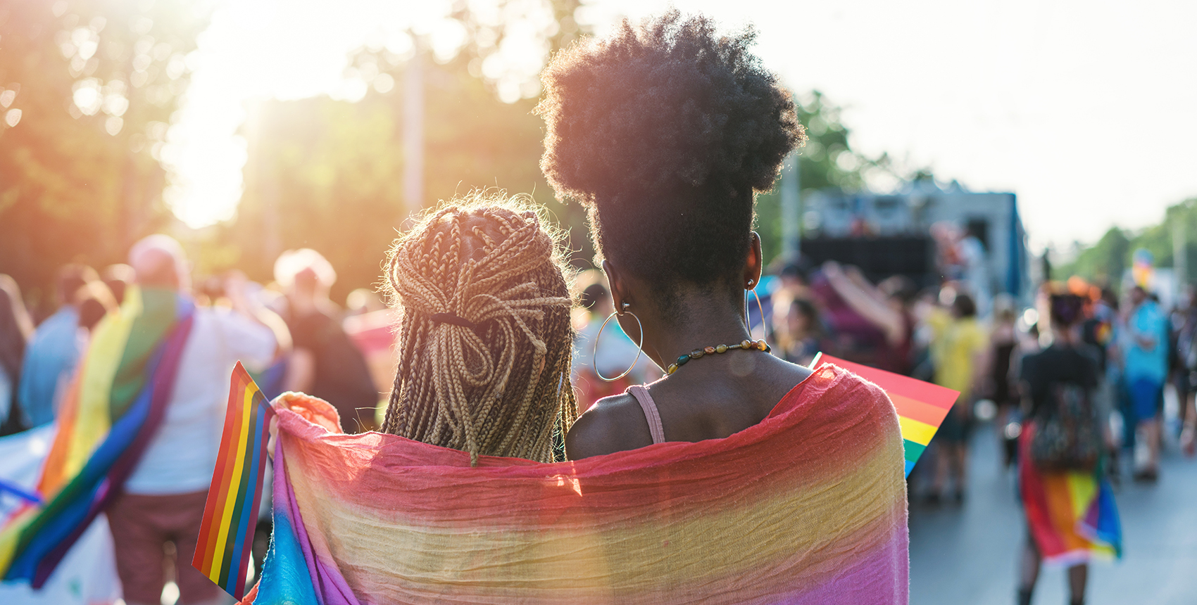 Rear view image of two people with braided, long hair, wrapped in a rainbow scarf walking with other people waving rainbow flags 