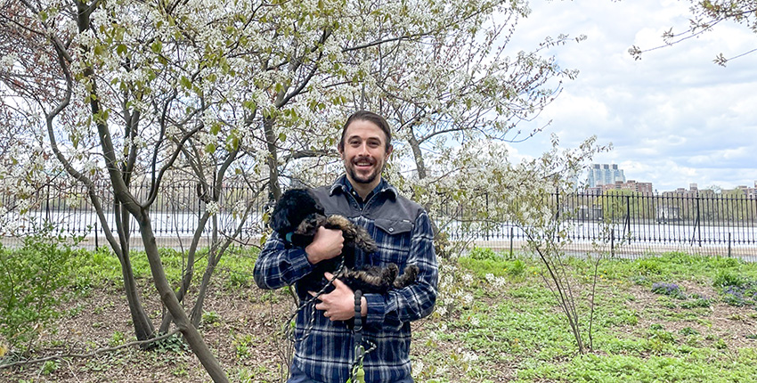 Nelson LaMarche with his miniature poodle in a NY city park with blooming pear tree and Hudson River behind him