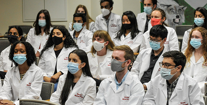 17 students wearing masks and in white coats that say Harvard Medical School seated in auditorium seats