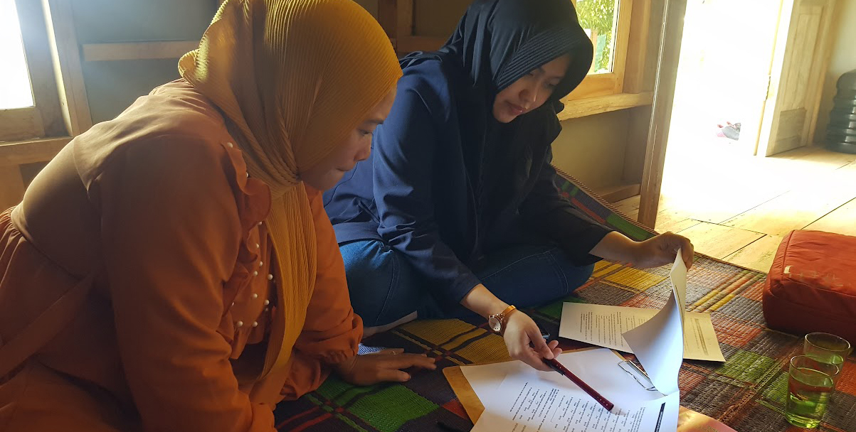 Two women, one in orange and one in blue seated on a colorful rug look at documents together.