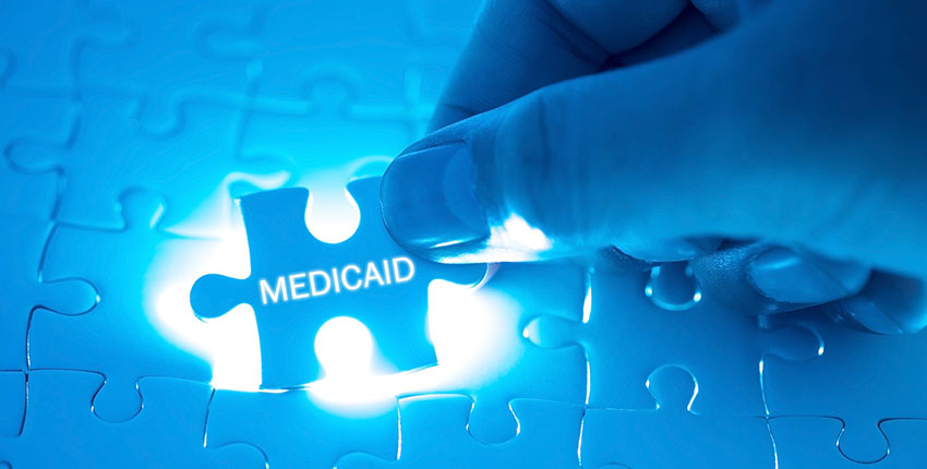A hand fitting a puzzle piece adorned with the word "medicaid" into a back-lit jigsaw puzzle.