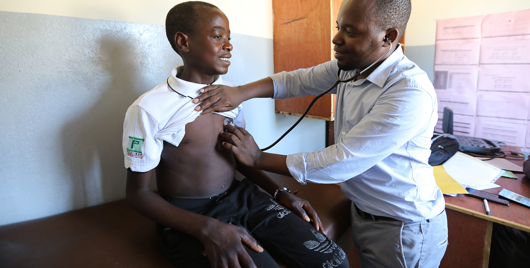 A teenage boy sits on an examination table in a clinic as a man listens to his heart with a stethoscope.