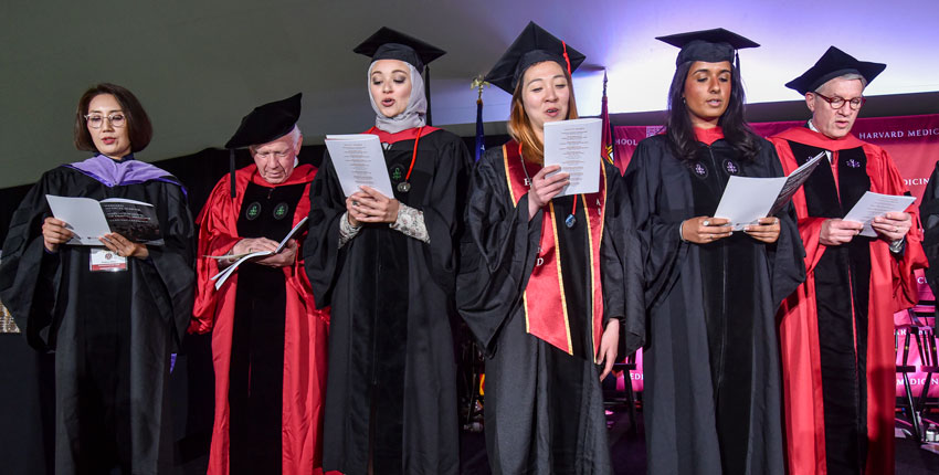 Group of six students and deans on stage holding programs and leading the audience in the reading of the oath.