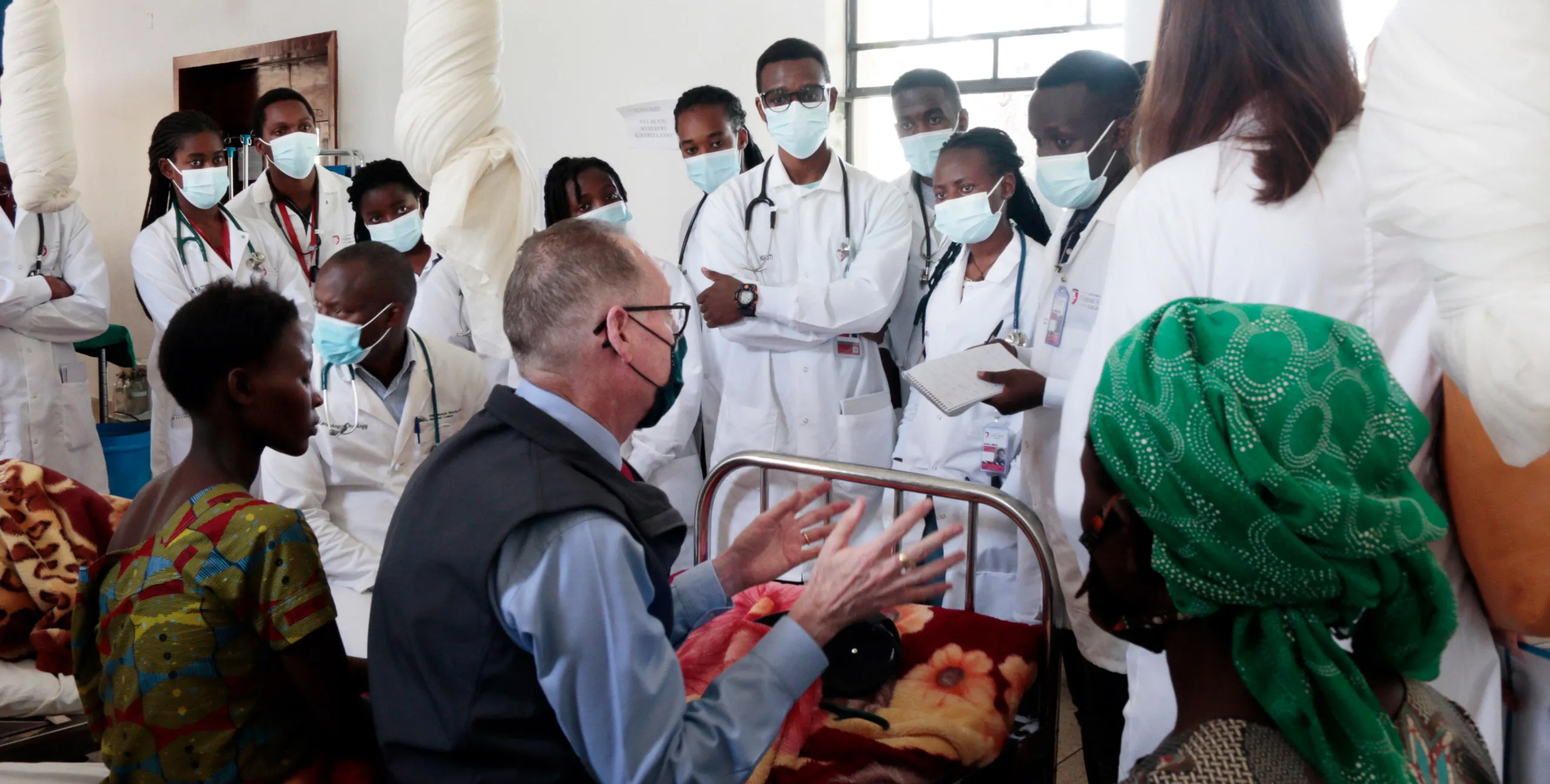 A line of women and men wearing white lab coats, medical masks, and stethoscopes stand facing three people, including Paul Farmer, sitting on hospital beds. 