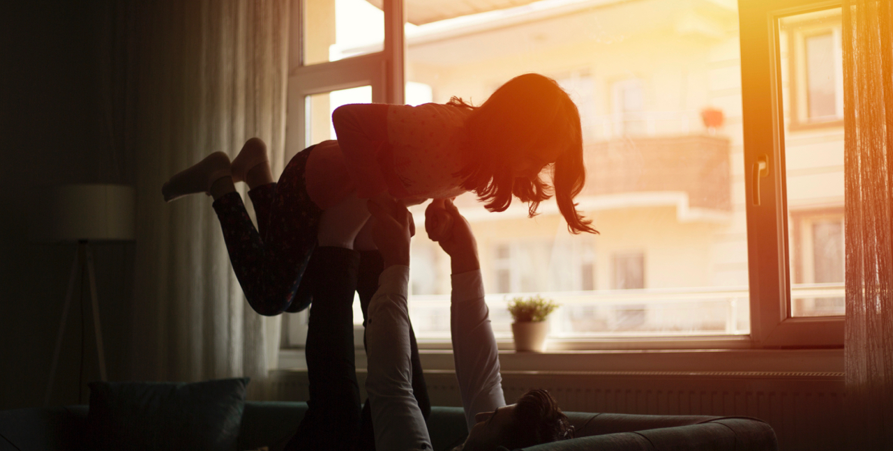 A father plays with his daughter backlit by sunset, seen through the window.