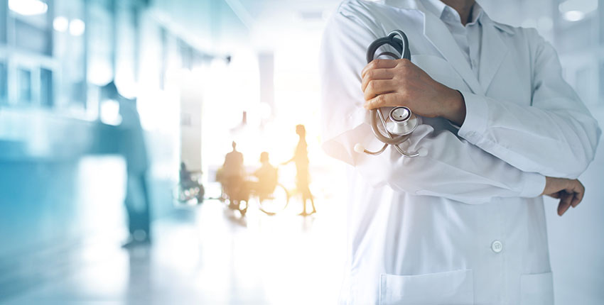 A physician with arms crossed stands in front of a blurry, overexposed background of a mostly empty waiting room.