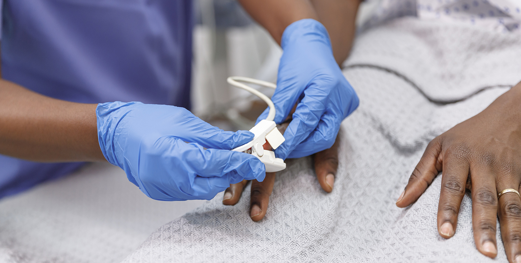 Close up shot of a medical professional placing a pulse oximeter on the finger of a Black hospitalized patient who is lying in bed.