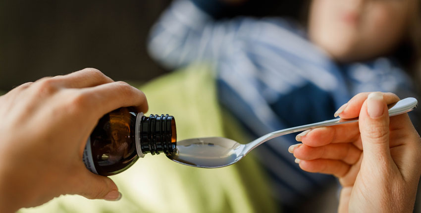 Closeup of adult women's hands pouring cough medicine into a spoon with child blurred out in background